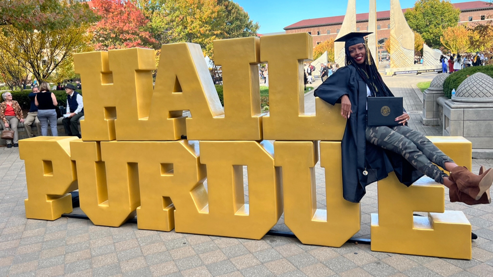 Gigi with her diploma, sitting on a yellow sign with letters saying Hail Purdue.