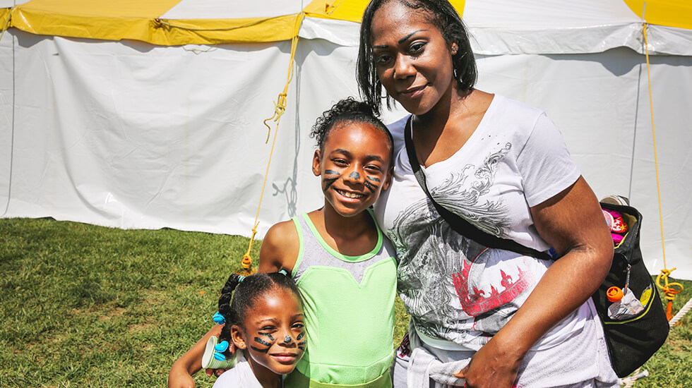 African American family, mother, and two daughters smiling in the outdoors outside of a fair tent