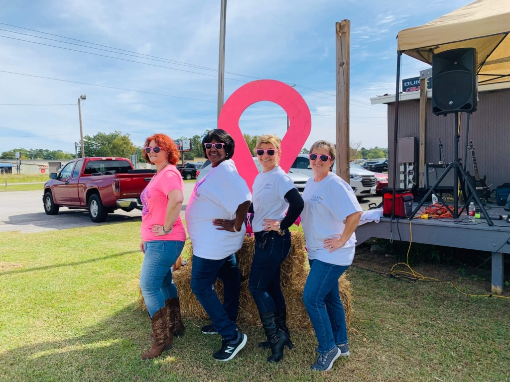 Sheri and volunteer team posing for the picture with a big pink ribbon behind them all wearing pink eyeglasses